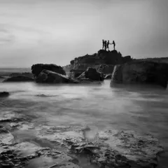 A long exposure shot turns the moving sea into a soft, misty surface, contrasting with the sharp silhouettes of people on the rocks.