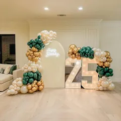 A chic and modern setup for a 25th birthday. This decoration features organic balloon arrangements in emerald green, gold, and cream, an arched backdrop with a neon sign, and large marquee numbers. It's a stylish way to celebrate a quarter-century.