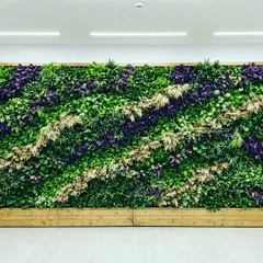 This artificial green wall features a striking diagonal pattern, using purple and light-colored foliage to create contrast against the green. The wooden base adds a natural finishing touch.