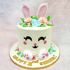 We craft pure pastel joy with this adorable bunny face cake. Perfect for a second birthday, this design features a sweet bunny face with large ears peeking out from a crown of delicate, handcrafted sugar flowers in soft peach and blue.