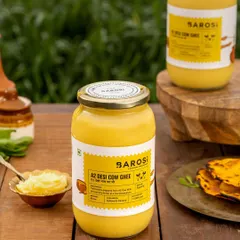 A lifestyle shot of Barosi's A2 Desi Cow Ghee, placed on a wooden table next to a stack of makki di roti. The outdoor, natural lighting and rustic props connect the product to traditional, wholesome Indian meals.