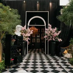 The entrance to a glamorous pre-wedding party. A black-and-white checkered path leads through a modern arched doorway into a room filled with art and elegance, flanked by lush cherry blossom arrangements.
