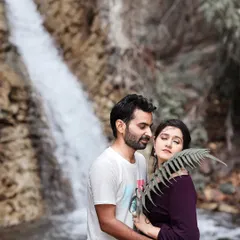 An intimate and artistic portrait by a waterfall. The use of a fern leaf adds a soft, natural element to the foreground, drawing focus to the couple's tender moment.