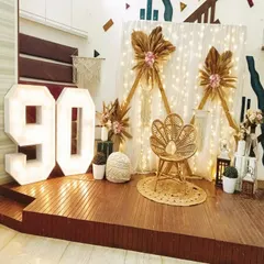 A rustic and chic 90th birthday setup on a wooden stage, featuring a peacock chair, macrame details, dried floral arrangements, and large marquee numbers.
