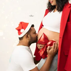 A heartwarming and creative Christmas shot. The husband-to-be kisses his wife's belly, which is adorned with a big red bow like the most precious gift.