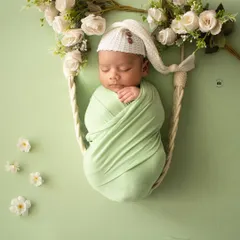 A sleeping newborn in a mint green wrap, nestled in a hanging swing prop adorned with white flowers. A dreamy and classic newborn portrait.