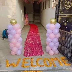 A beautiful welcome entrance for a baby girl's welcome home party. The decor features a path of pink rose petals, the word 'WELCOME' written with marigold flowers, and two balloon pillars in pink and purple.