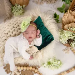 A beautiful boho setup featuring a baby in a white knit outfit sleeping on a chunky wool blanket. The scene is decorated with wicker baskets, dried palm leaves, and green foliage.