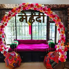 For this home Mehndi function, I created a beautiful and vibrant seating area right by the window. A bright pink sofa is framed by a colorful floral arch and the word "Mehndi" in Hindi script. The setup is completed with traditional Rajasthani umbrellas, making it a perfect photo spot.