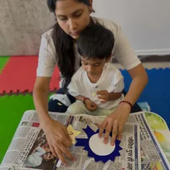 A mother helps her child paste shapes onto a paper medal, a craft that builds fine motor skills and gives them a sense of accomplishment. We believe in creating tangible results that children can be proud of.