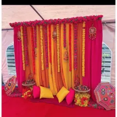 A festive Mehendi seating area with a backdrop of pink, orange, and yellow drapes. The decor includes traditional hangings, Rajasthani umbrellas, and comfortable floor seating with cushions.