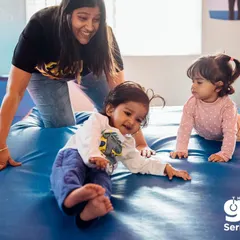 An instructor smiles as she guides two toddlers in a gentle crawling exercise on a soft mat. This foundational movement helps build core strength and coordination, preparing them for more complex skills later on.