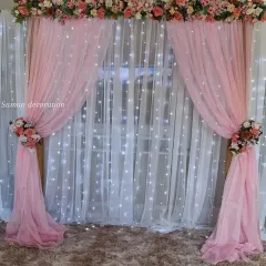 A beautiful photo booth backdrop made with a curtain of white fairy lights. The pink fabric drapes and floral arrangements add a touch of elegance.