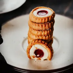A close-up shot of jam-filled cookies stacked in a tower. The moody lighting and tight focus on the bite taken out of the front cookie create a sense of indulgence and rustic charm.