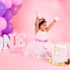 A wide shot of a celebratory first birthday cakesmash, with the baby girl in a pink dress, a lit-up "ONE" sign, and a beautiful cake.