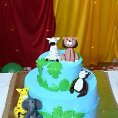 A delightful two-tier jungle-themed cake. It features cute, edible animal figures like a zebra, lion, and monkey, perfectly matching the party's theme.