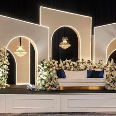 A modern and sophisticated reception stage. The design features clean white geometric arches with built-in lighting, crystal chandeliers, and elegant white floral decor.