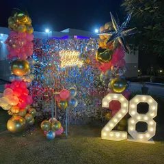 An outdoor shimmer wall setup for a 28th birthday. The pink, gold, and silver balloons, along with the neon sign and marquee numbers, create a festive party vibe.