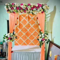 A full wall backdrop created with a lattice of orange and white artificial flowers. This textured and vibrant background is framed by peach-colored drapes, offering a unique and eye-catching setting for Bappa.