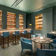 This angle focuses on the relationship between the bar and the dining area at Armani Caffe. The composition highlights the well-stocked, warmly lit bar as a focal point, while showing its connection to the intimate table settings, perfect for restaurant interior photography.