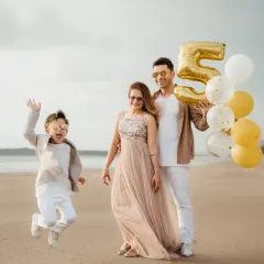 A young boy jumps for joy during his fifth birthday photoshoot on the beach. His parents look on, holding balloons, making for a perfect family portrait.