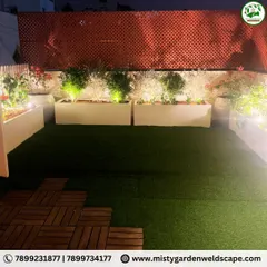 A beautiful night view of a transformed terrace garden. The artificial lawn is surrounded by illuminated planters and a PVC lattice screen for privacy, creating a magical evening ambiance.