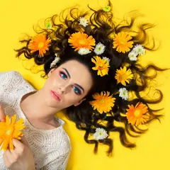 An overhead beauty shot of a model lying on a bright yellow background with yellow and white flowers scattered through her hair. This high-contrast, colorful image is perfect for a hair care or beauty campaign.