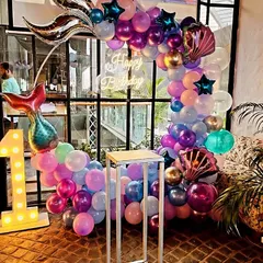An underwater fantasy with this mermaid-themed first birthday party. The decoration includes a balloon arch in purple and blue, mermaid tail foils, and a large light-up number '1'.