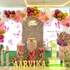 A first birthday celebration with a pink and gold butterfly theme. The setup includes a shimmer wall, a large light-up number '1', and a cute fairy cutout.