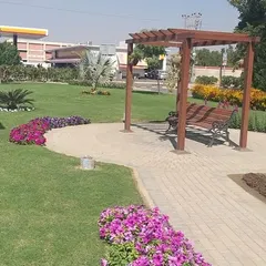 A different view of the public park, showing the seating area with a pergola, surrounded by colorful flower beds and a healthy lawn. This is a space designed for community relaxation and enjoyment.