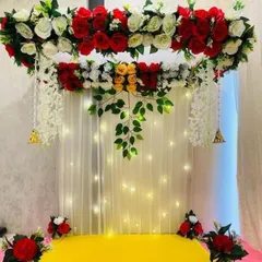 This backdrop features a beautiful arch of red, white, and yellow roses over a curtain of warm fairy lights. The design includes hanging white floral strings and bells, blending modern style with traditional elements.