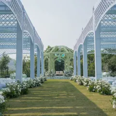 The long aisle leading to the green mandap, flanked by white lattice archways that create a beautiful, structured pathway for the wedding procession.