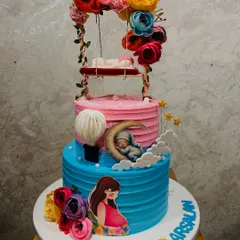 A beautiful two-tier baby shower cake in pink and blue. The design features a sleeping baby on a floral swing, a pregnant mother illustration, and a hot air balloon.