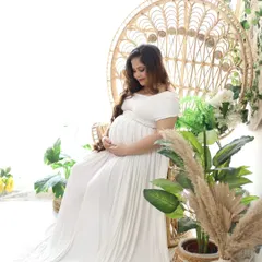 A serene portrait of a mom-to-be, lost in thought while cradling her bump. The white gown and natural props like potted plants and a peacock chair create a peaceful atmosphere.