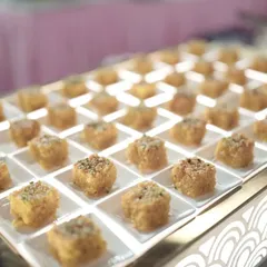 Bite-sized squares of a traditional Indian sweet, possibly a milk-based barfi, are presented on an illuminated tray. The grid-like arrangement creates a clean, contemporary look for the dessert counter.