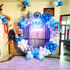 A Boss Baby themed birthday decoration in a home setting. The circular backdrop is framed with blue, white, and silver balloons and features a shiny fringe curtain and a neon sign.