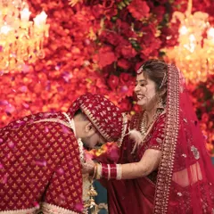Bowing down for love is the best win. This playful moment during the jaimala ceremony, against a stunning red floral backdrop, is full of joy and adoration.