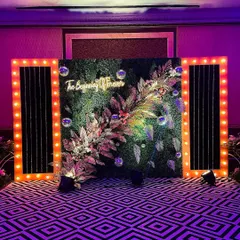 "The Beginning of Forever." This unique photobooth combines a lush green wall with modern bulb-lit frames and a custom neon sign, creating a stylish and meaningful backdrop.