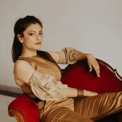 A stylish indoor portrait featuring a vintage red chair as a prop. The contrast between the neutral outfit and the bold furniture creates a visually interesting and fashionable shot.