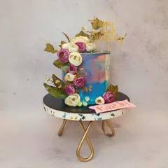 A single-tier cake with a beautiful watercolor buttercream effect in shades of blue and purple. A lush cascade of artificial flowers in complementary colors makes this a truly artistic and blooming creation.