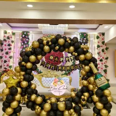 An elegant and grand retirement party decoration in a banquet hall. The setup features a large circular arch with classic black and gold balloons, a 'Happy Retirement' banner, and a champagne bottle foil balloon for a celebratory touch.