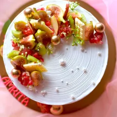 A beautiful fresh fruit cake, topped with a mix of seasonal fruits like figs, kiwi, and pomegranate. The fruit is arranged artfully on a base of white cream with a textured finish.