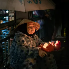 A poignant portrait of an elderly Vietnamese woman in a traditional conical hat, selling candle-lit lanterns from a boat at night.