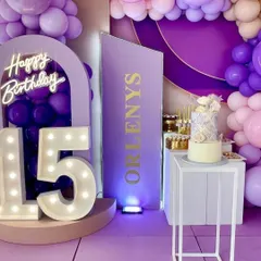 A stylish 15th birthday party with a purple and lavender color scheme. The setup includes marquee numbers, a custom name sign, and a modern cake display.