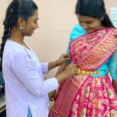 A student helps her partner drape a half saree, securing it with a traditional waistband. These practical sessions are where real learning happens.
