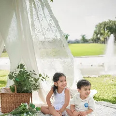 Two siblings sit together on a picnic blanket, smiling and enjoying the beautiful day. The fountain in the background adds a lovely touch to the scene.