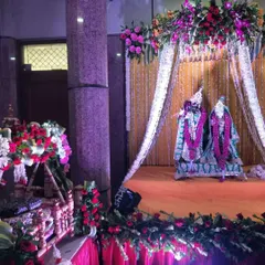 A complete Janmashtami scene with idols of Radha and Krishna under a beautifully decorated canopy. A small jhula for Ladoo Gopal is placed to the side, surrounded by fresh flower arrangements.