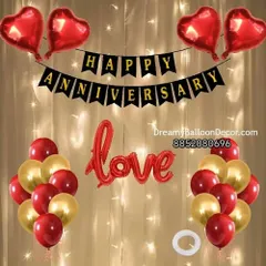 A classic anniversary decoration kit. This setup includes a 'Happy Anniversary' banner, 'Love' foil balloon, and bunches of red and gold balloons, all set against a curtain of warm fairy lights.
