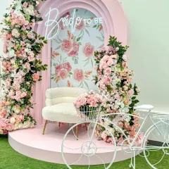 A charming "Bride to be" photo corner with a pink arched backdrop and a floral-print interior. The setup is completed with a plush white chair, a decorative white bicycle, and an abundance of pink roses.