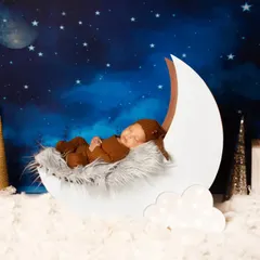 A newborn in a cozy brown outfit sleeps peacefully inside a white crescent moon prop. This setup uses a softer, fluffier texture for the moon and clouds.